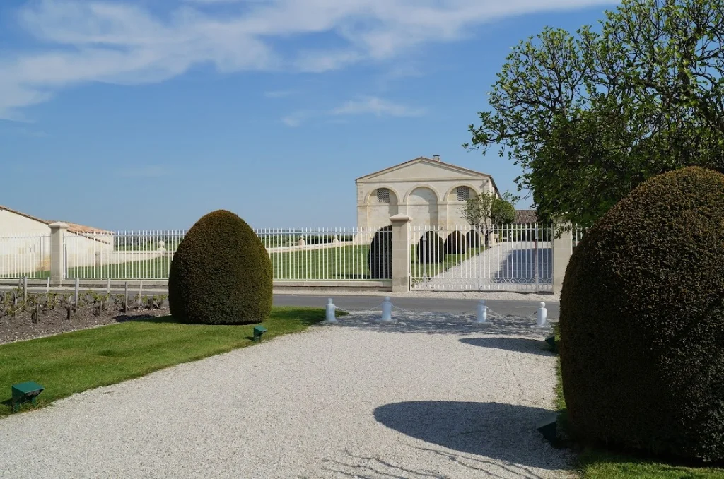 2013-04-29に酒井 浩（プラチナワイン（有限会社デリバリー・ワイン）代表）がシャトー・ムートン・ロートシルト / Pauillac, Médoc, Bordeaux, Franceで撮影した離れた位置からシャトー・ムートン・ロートシルトのパヴィリオン正門方向を望む遠景。左右を黒い鉄柵で仕切られたグラベルアプローチと、奥に建つ新古典主義建築が確認でき