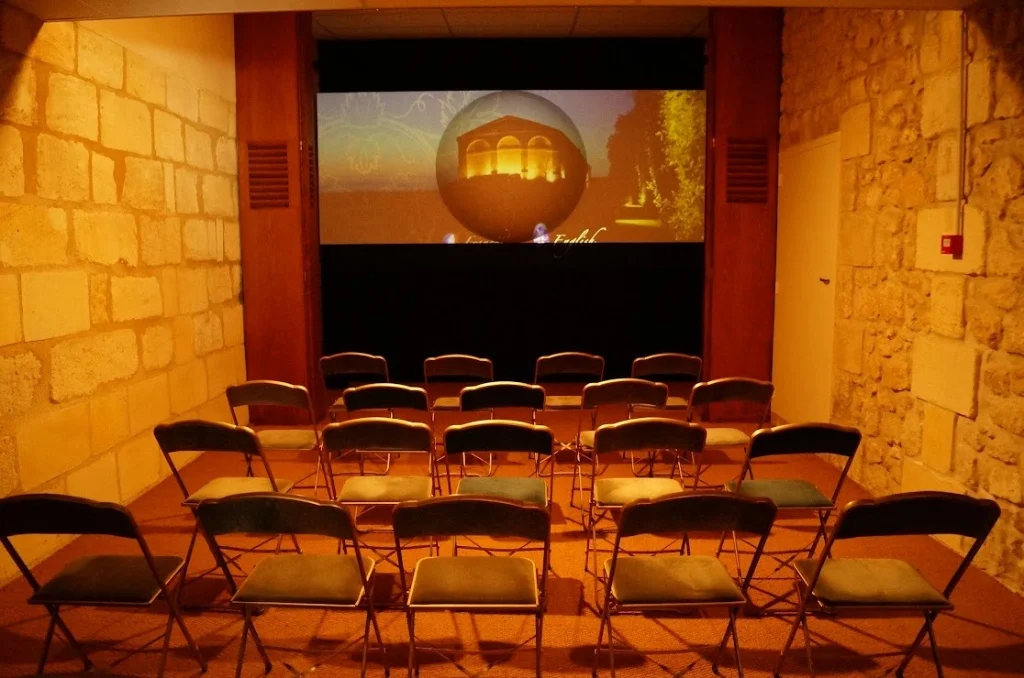 2013-04-29に酒井 浩（プラチナワイン（有限会社デリバリー・ワイン）代表）がシャトー・ムートン・ロートシルト / Pauillac, Médoc, Bordeaux, Franceで撮影したシャトー見学の冒頭で上映されるムートンの歴史を紹介するフィルム上映室。石灰岩の石壁に囲まれた重厚な空間に折りたたみ椅子が並ぶ。1973年の一級昇格やアーティスト